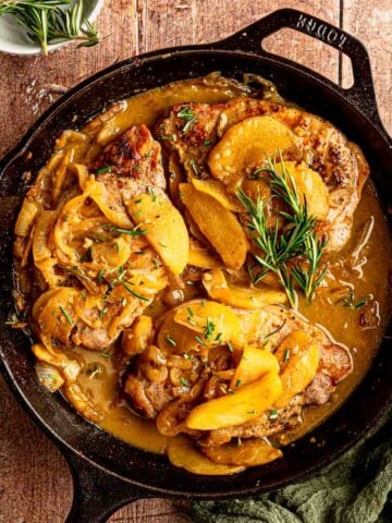 Cooked pork chops nestled in a skillet with caramelized apples, onions, cider sauce, and fresh rosemary.