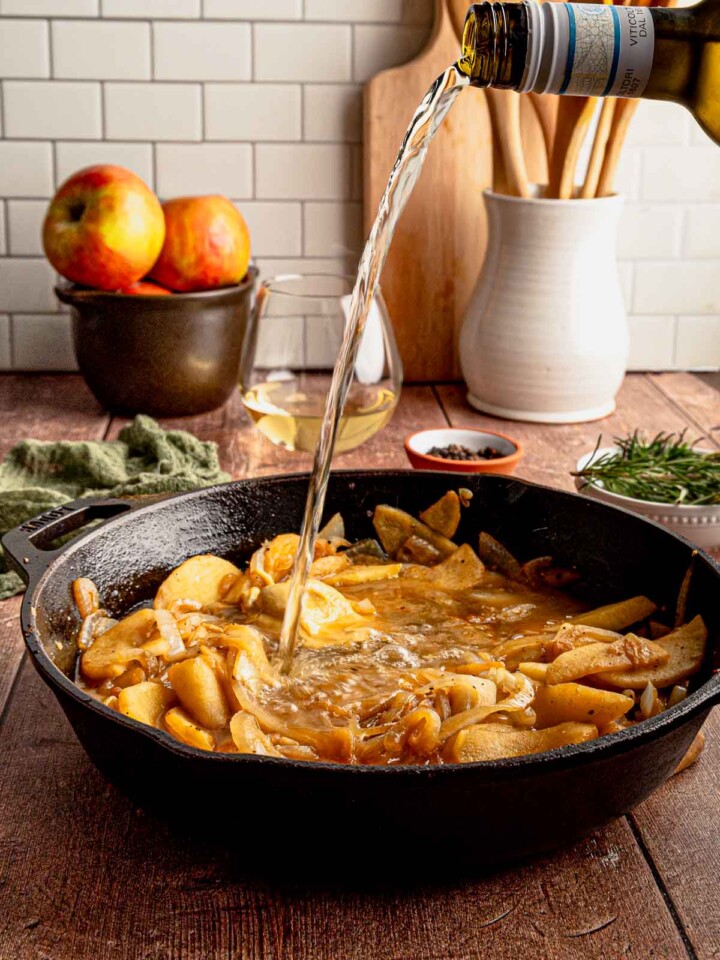 A bottle of white wine pouring into a skillet of apples, onions, and mustard for a flavorful pan sauce.