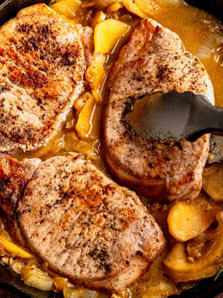 Juicy pork chops simmering in a rich apple cider sauce with onions and apples in a skillet.