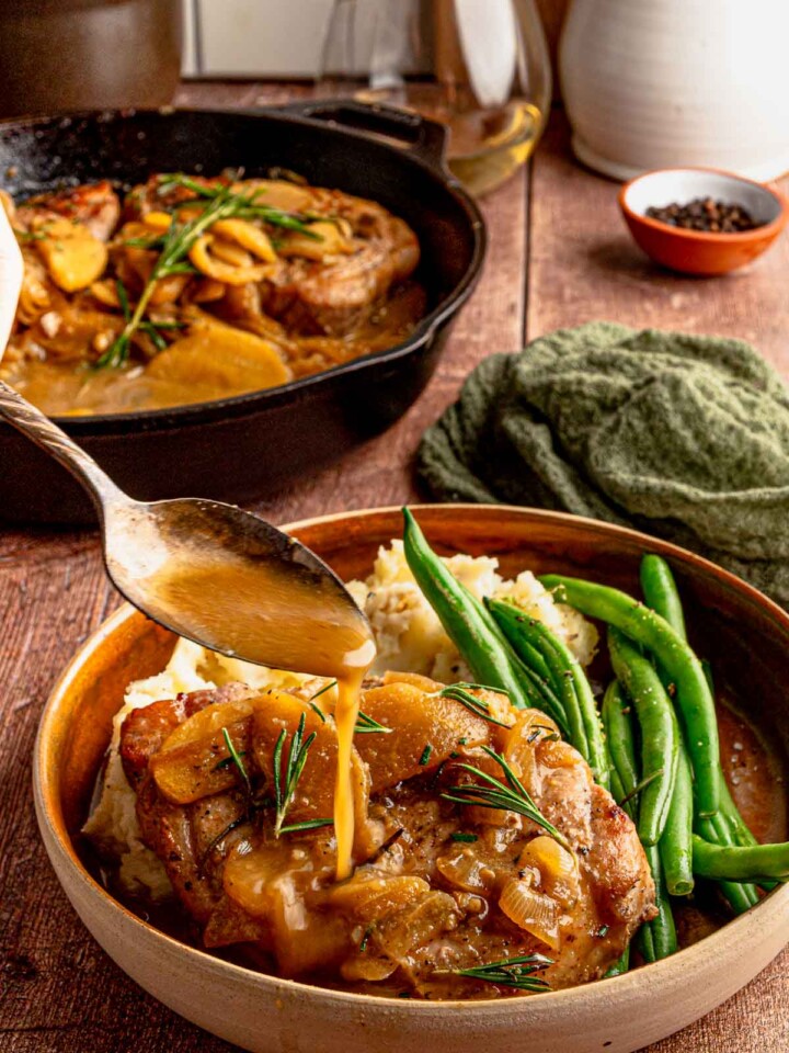 A spoon drizzling apple cider sauce over pork chops served with mashed potatoes and green beans.