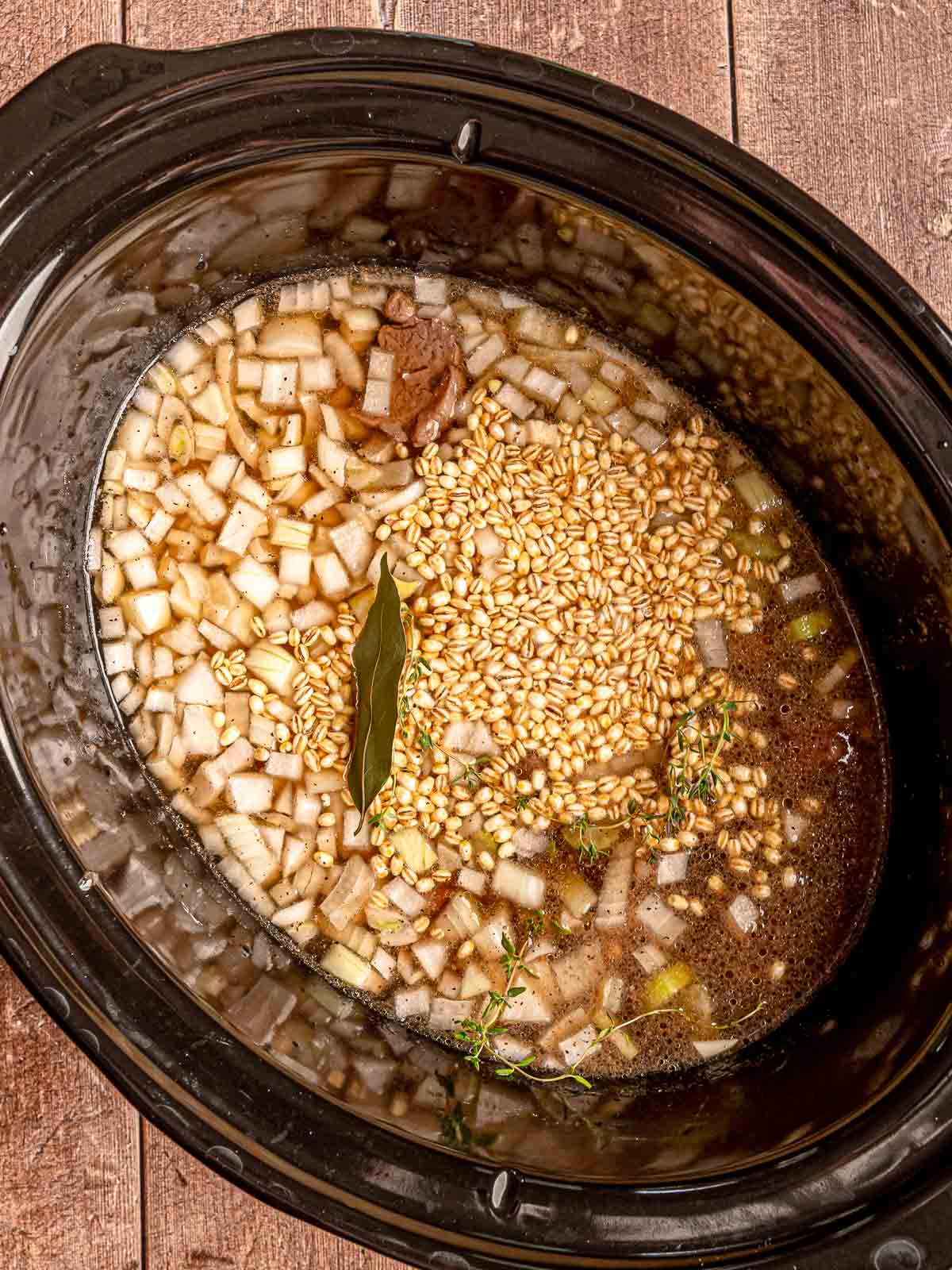 Beef, barley, onions, broth, bay leaf, and thyme in a slow cooker before cooking.