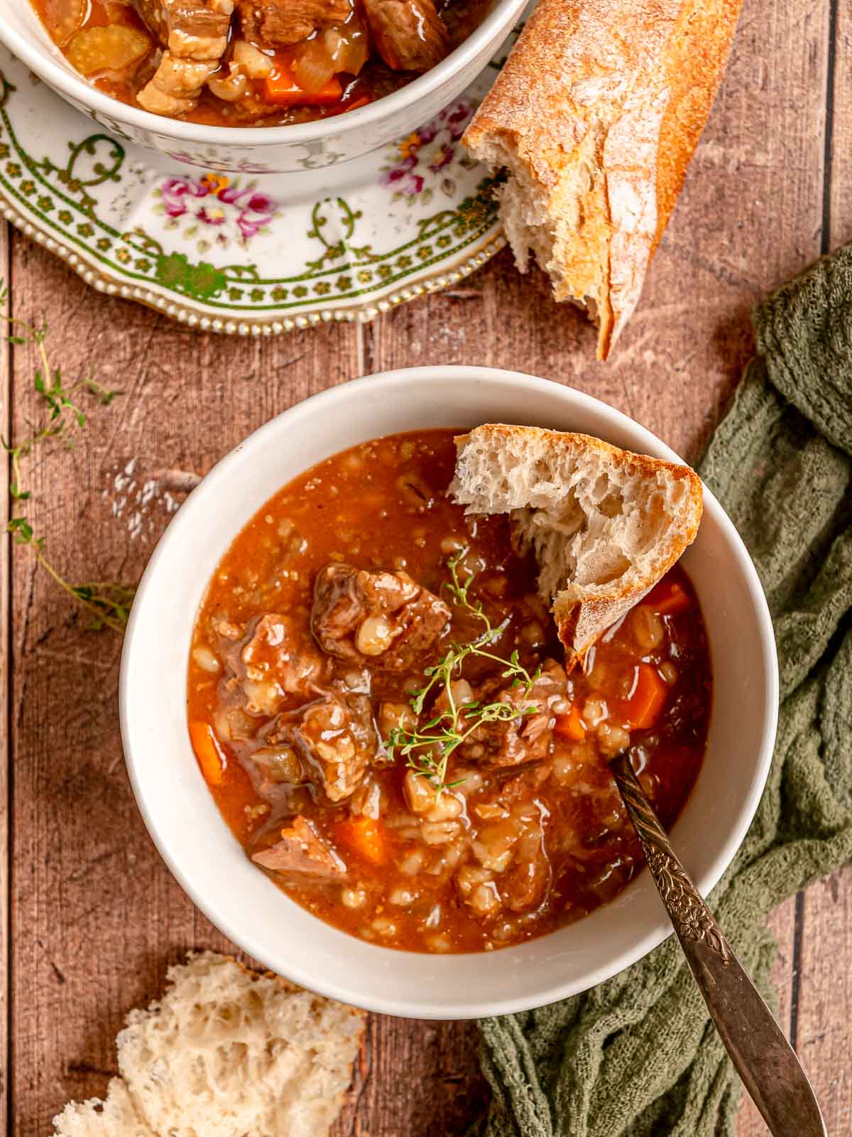 Beef and barley soup garnished with thyme, served in a white bowl with bread on the side.