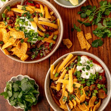 Two bowls of loaded beef chili with toppings of cheese, sour cream, corn chips, scallions, and cilantro on a wooden table.