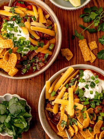 Two bowls of loaded beef chili with toppings of cheese, sour cream, corn chips, scallions, and cilantro on a wooden table.