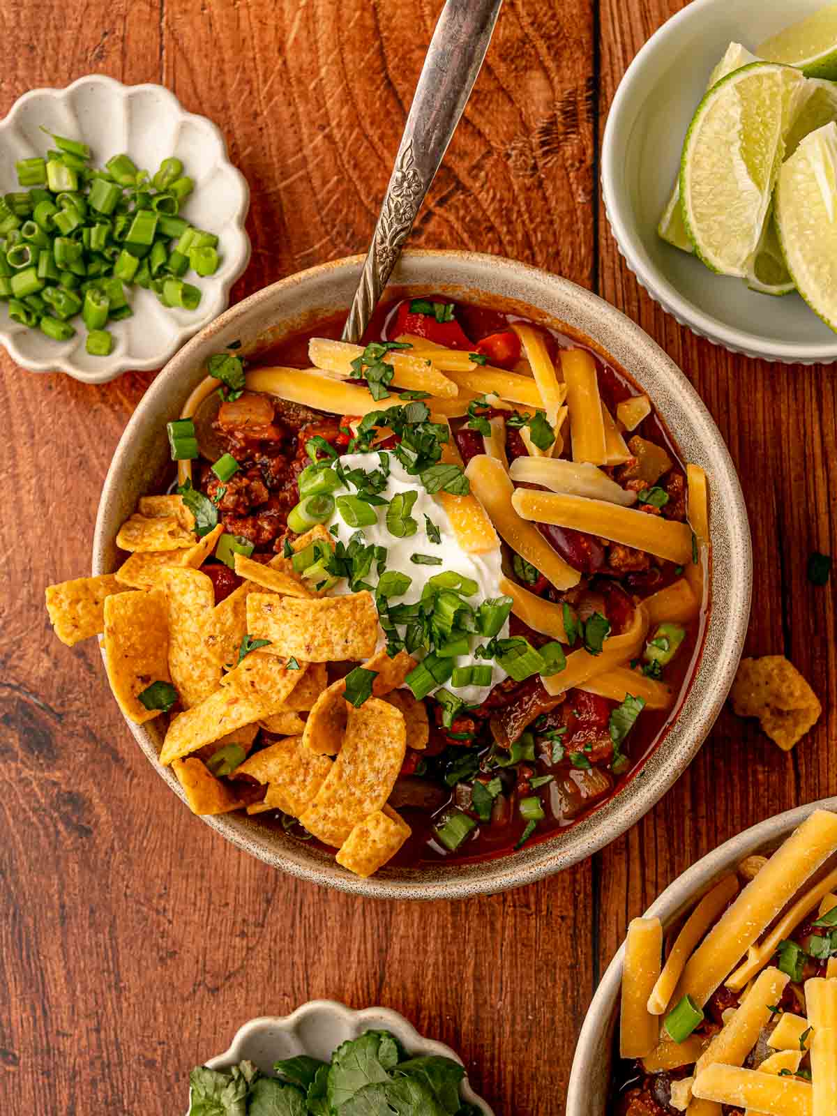 Bowl of hearty beef chili garnished with cheese, sour cream, and scallions, served with corn chips on top.