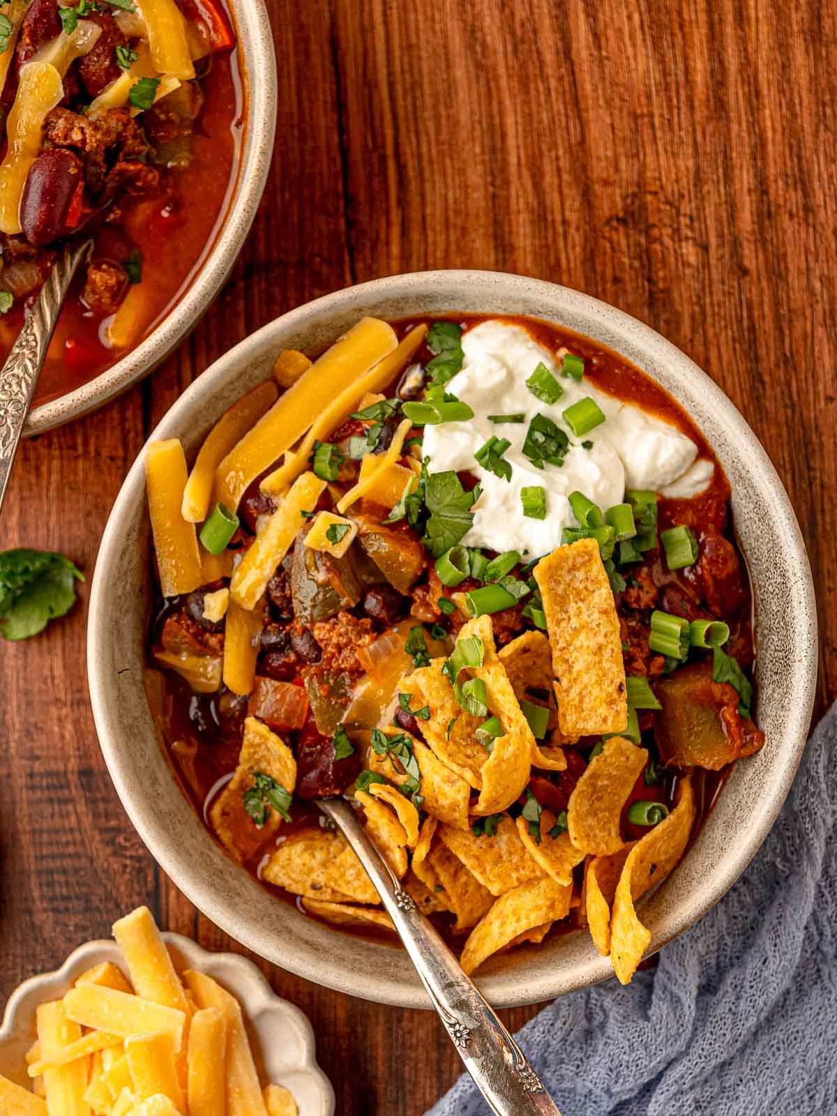 Bowl of beef chili topped with shredded cheddar cheese, sour cream, scallions, cilantro, and corn chips, with lime wedges on the side.