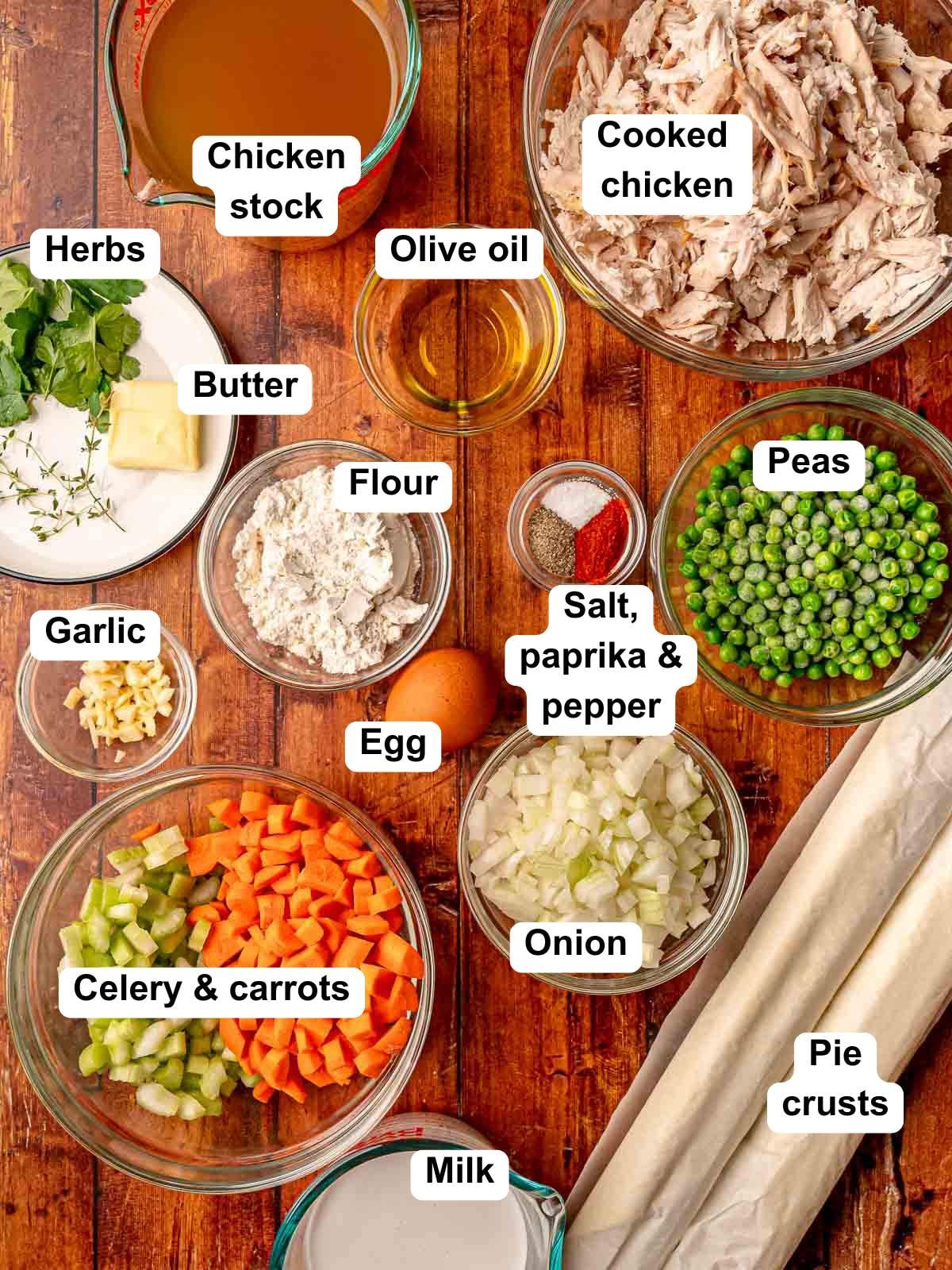 Ingredients for chicken pot pie arranged on a wooden surface including chicken, vegetables, pie crusts, and seasonings.