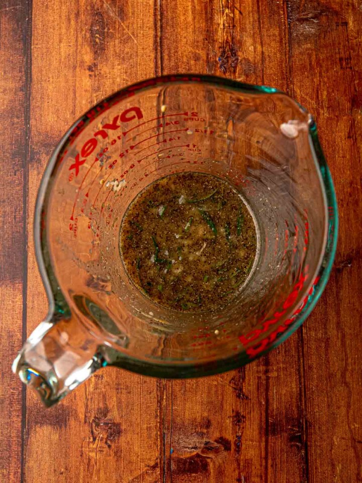 A glass measuring cup filled with the garlic rosemary maple marinade made with olive oil, lemon, Dijon mustard, and herbs.