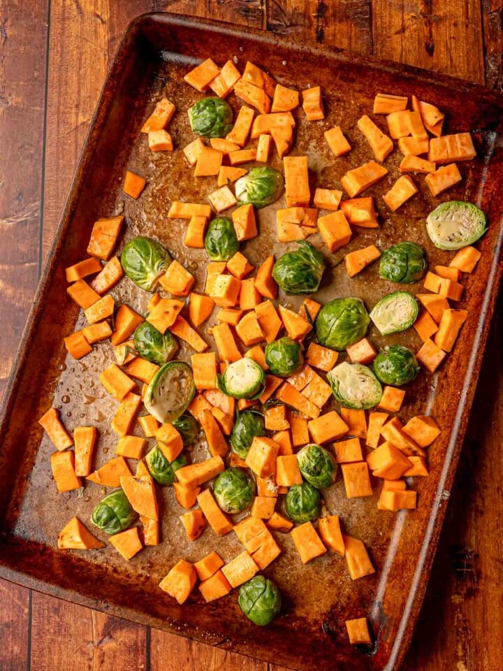 A baking sheet with chopped sweet potatoes and Brussels sprouts lightly coated in oil and seasoning, ready to roast.