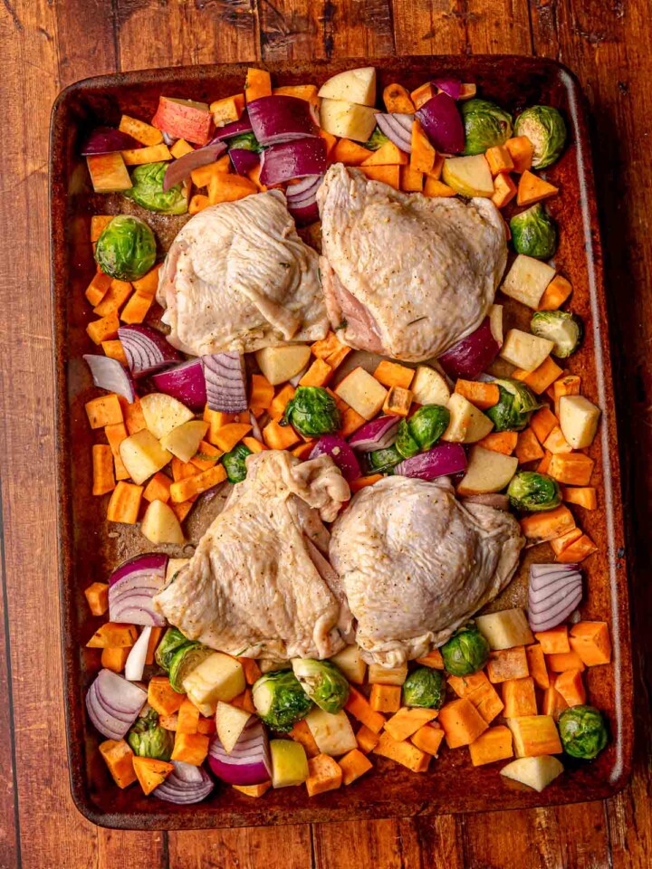 Chicken thighs placed on a sheet pan surrounded by seasoned sweet potatoes, Brussels sprouts, red onions, and apples before roasting.