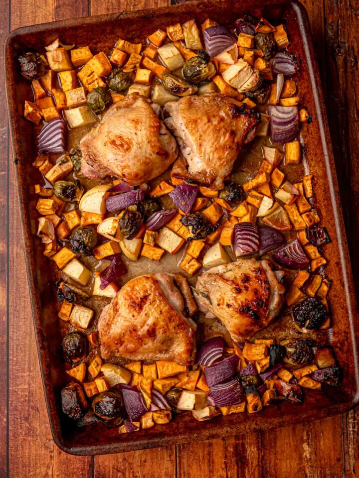 Chicken thighs placed on a sheet pan surrounded by seasoned sweet potatoes, Brussels sprouts, red onions, and apples before roasting.