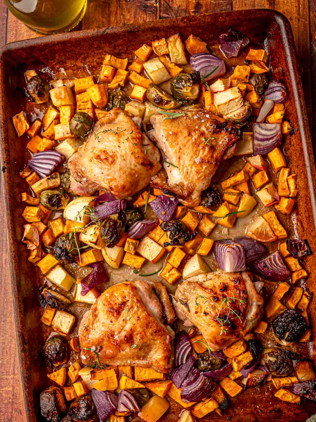 Crispy roasted chicken thighs surrounded by roasted apples, red onion, and vegetables with a drizzle of maple glaze and fresh herbs on top.