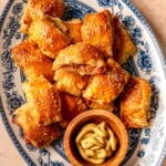 Golden chicken sausage rolls stacked on a floral platter next to a small bowl of mustard.