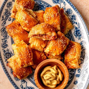 Golden chicken sausage rolls stacked on a floral platter next to a small bowl of mustard.