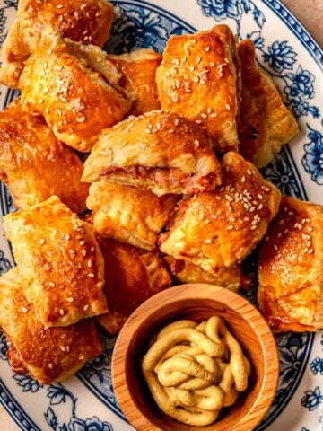 Golden chicken sausage rolls stacked on a floral platter next to a small bowl of mustard.