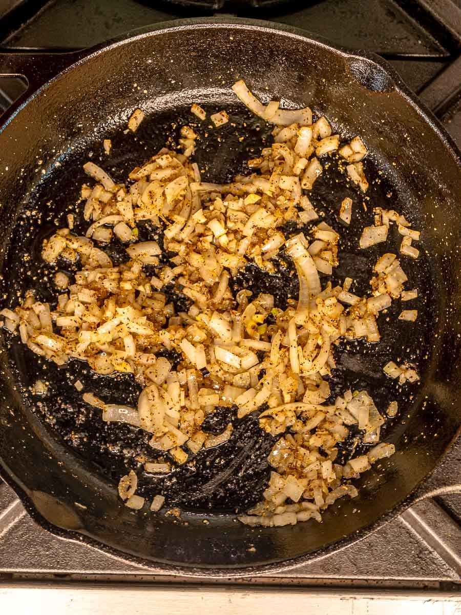 Diced onions and garlic sautéing with seasonings in a cast-iron skillet.