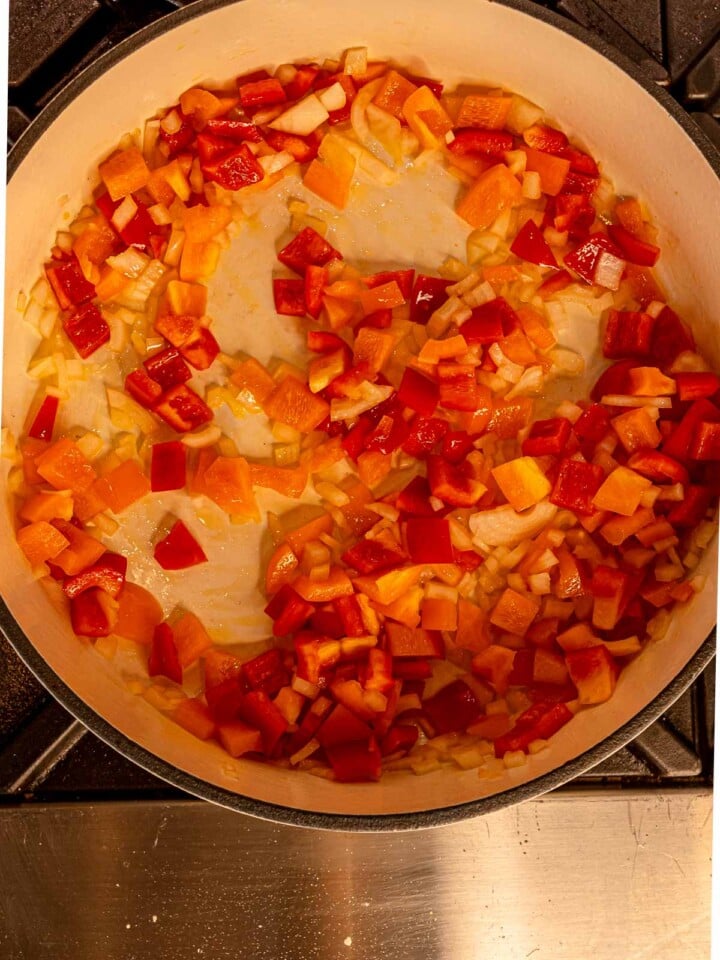 Diced red and orange bell peppers sautéing with onions in a large skillet.