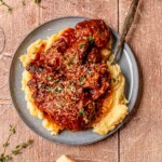 Italian pot roast served over mashed potatoes on a gray plate with gravy and herbs.