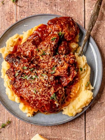 Italian pot roast served over mashed potatoes on a gray plate with gravy and herbs.