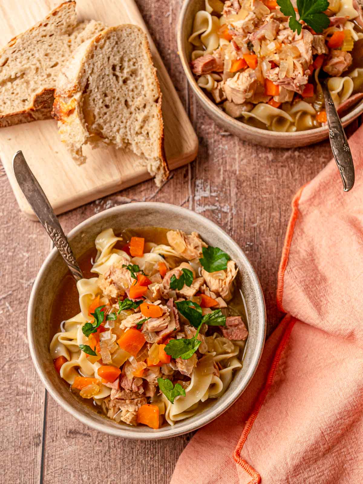 Two bowls of turkey noodle soup with egg noodles, bread slices, and a peach napkin.