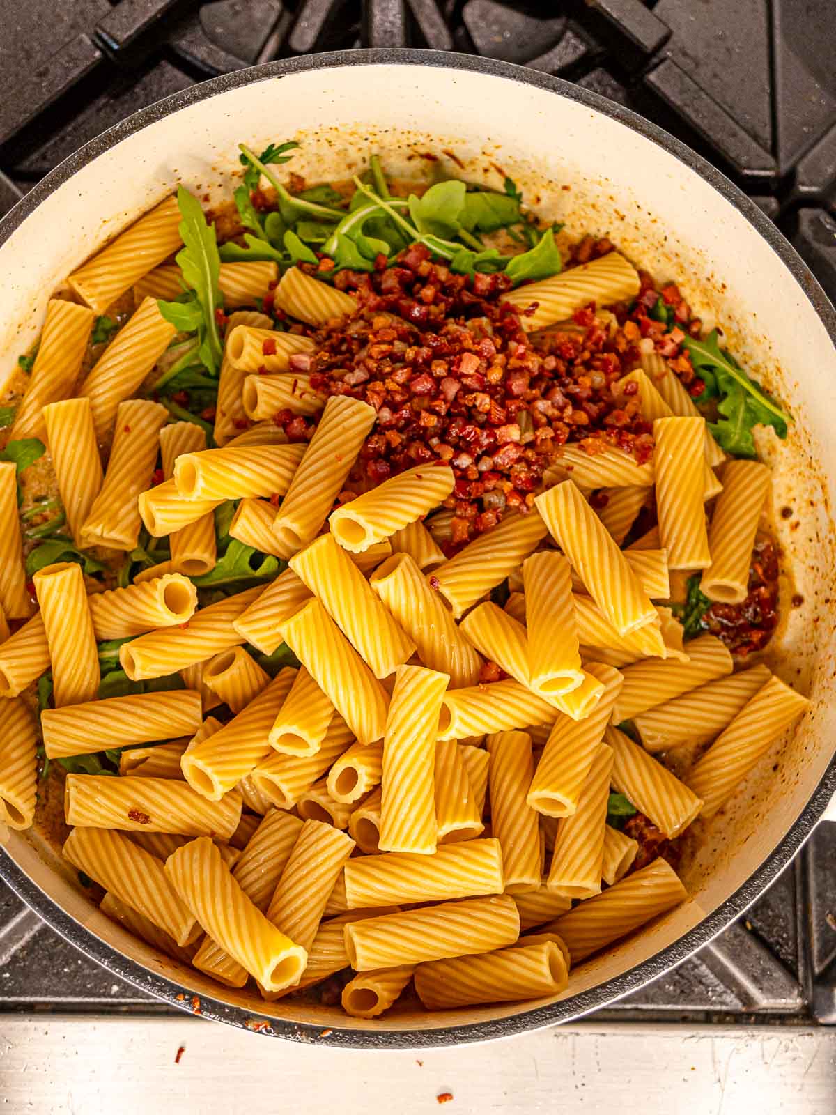 Pasta and cooked pancetta in a skillet with arugula and sauce.