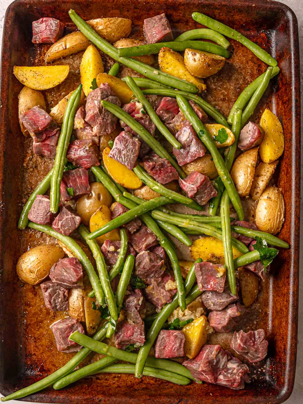 Raw sirloin steak bites, potatoes, and green beans arranged on a sheet pan with garlic-butter seasoning.