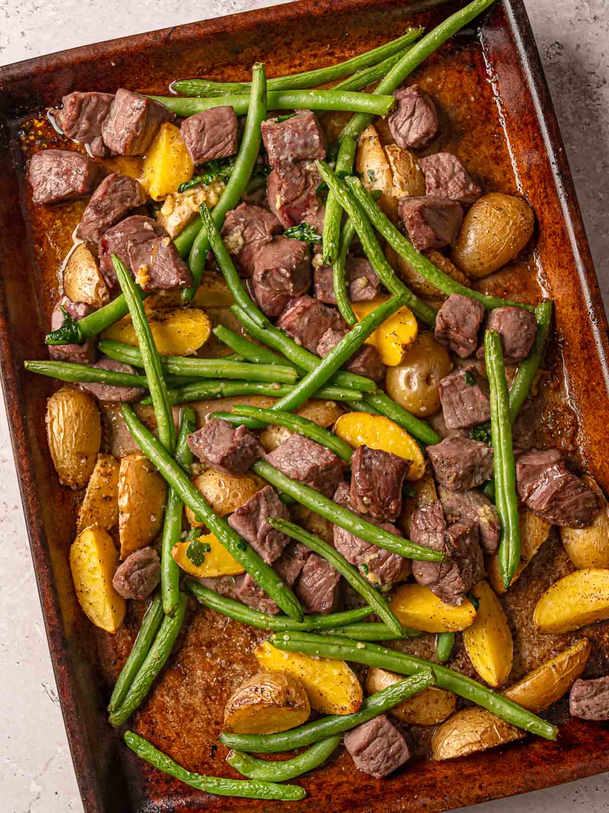 Sirloin steak bites, green beans, and potatoes on a sheet pan coated in garlic-butter seasoning.