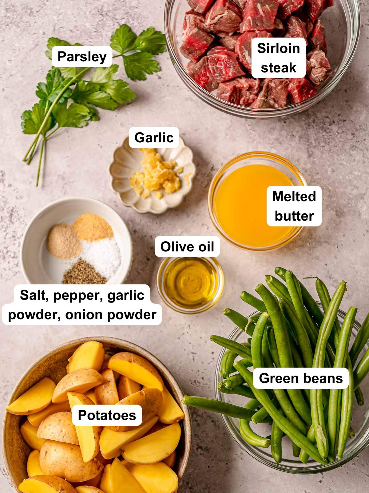 Sirloin steak bites, potatoes, green beans, melted butter, garlic, olive oil, dried seasonings, and parsley displayed on a counter.