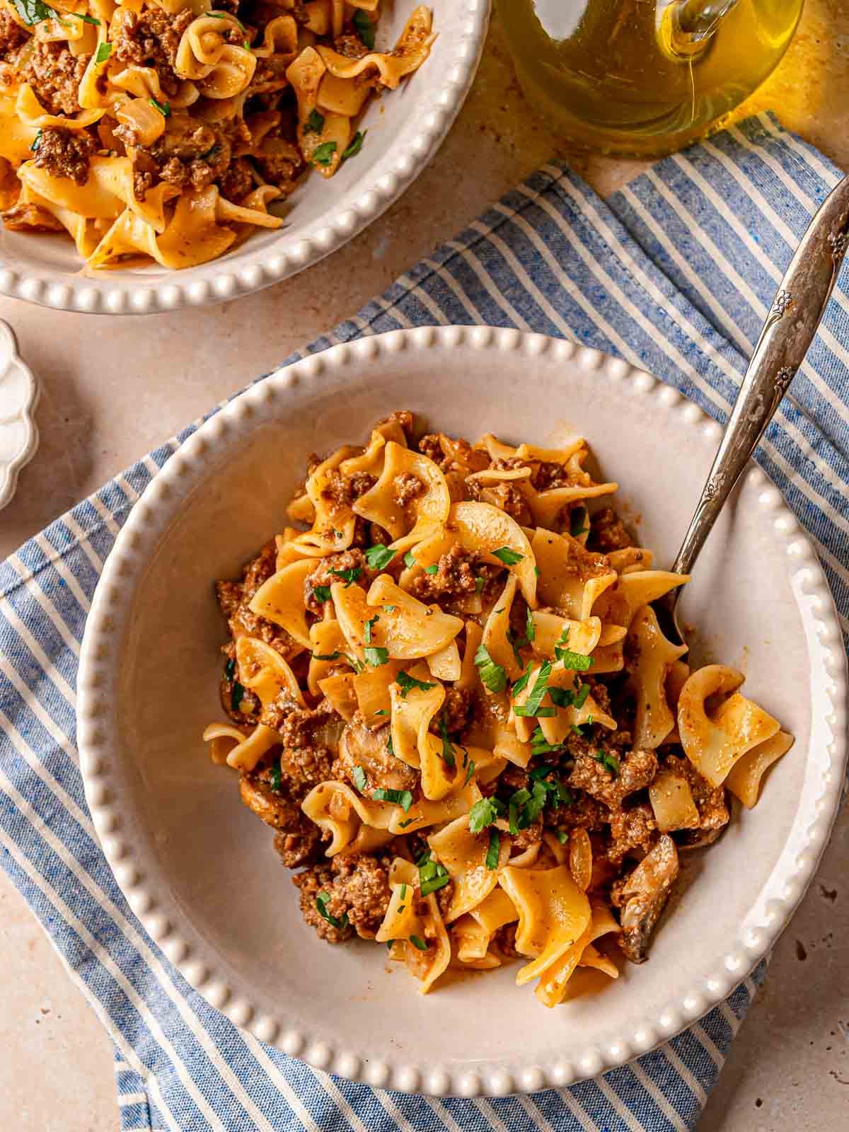 Two bowls of ground beef stroganoff with egg noodles, garnished with parsley and ready to serve.