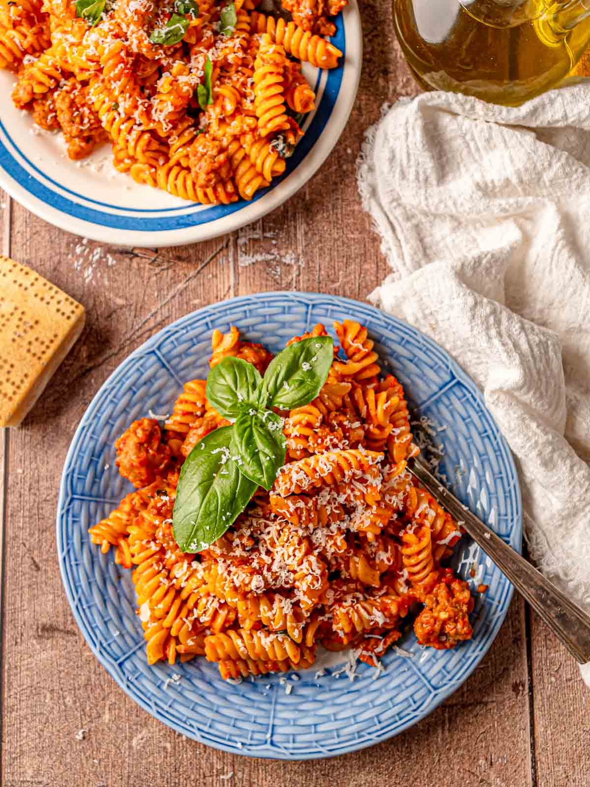 Roasted red pepper pasta with sausage served on a blue plate with grated Parmesan and basil.