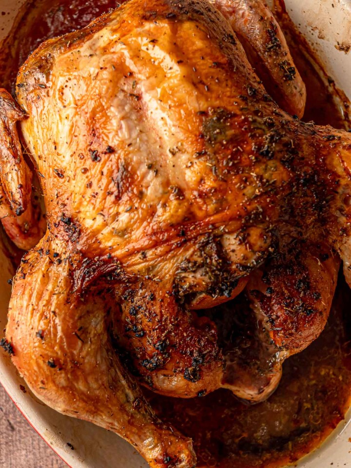 A close-up, top-down shot of a golden-brown roasted chicken with crispy skin and visible dried herbs in a roasting pan.