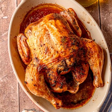 A whole roasted chicken with deep golden-brown skin, resting in its juices inside a ceramic baking dish.