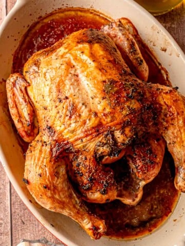 A whole roasted chicken with deep golden-brown skin, resting in its juices inside a ceramic baking dish.