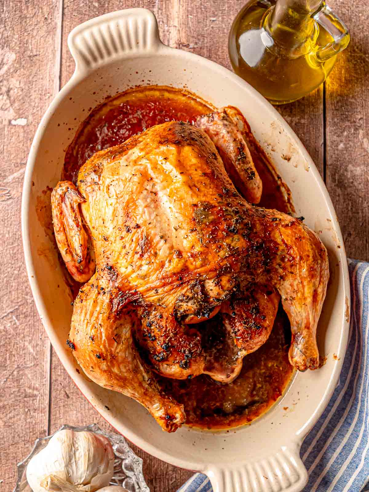 A finished Greek roasted chicken in a baking dish, styled on a wooden surface with a bottle of olive oil, a head of garlic, and a blue striped napkin.