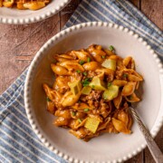 Cheeseburger pasta in a white scalloped-edge bowl with dill pickles and green onions on a blue striped napkin.