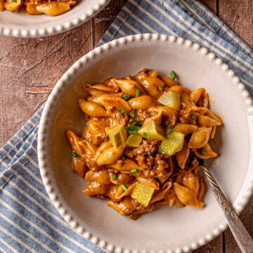 Cheeseburger pasta in a white scalloped-edge bowl with dill pickles and green onions on a blue striped napkin.