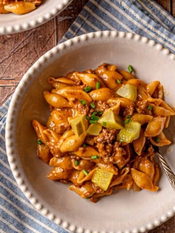 Cheeseburger pasta in a white scalloped-edge bowl with dill pickles and green onions on a blue striped napkin.