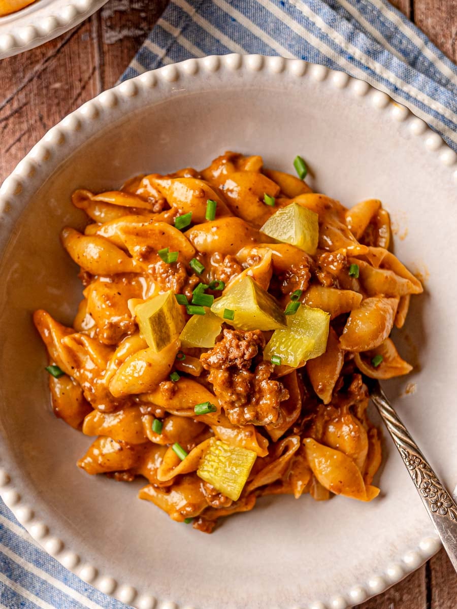 Cheeseburger pasta in a white scalloped-edge bowl, topped with dill pickles and green onions.
