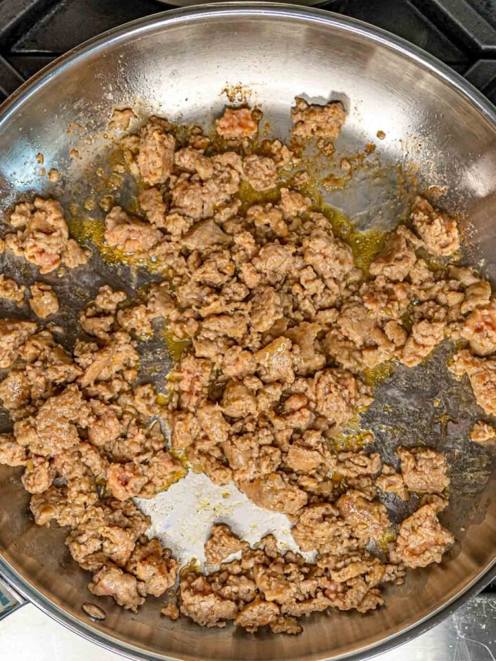 Crumbled Italian sausage browning in a stainless steel skillet with rendered fat.
