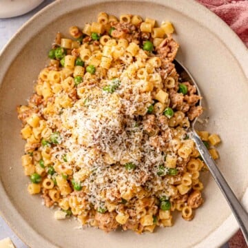 A stoneware bowl filled with ditalini pasta, crumbled sausage, and peas, finished with grated Parmesan and cracked black pepper, with a spoon and pink linen alongside.
