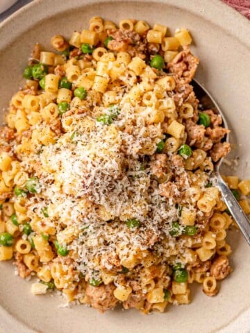 A stoneware bowl filled with ditalini pasta, crumbled sausage, and peas, finished with grated Parmesan and cracked black pepper, with a spoon and pink linen alongside.