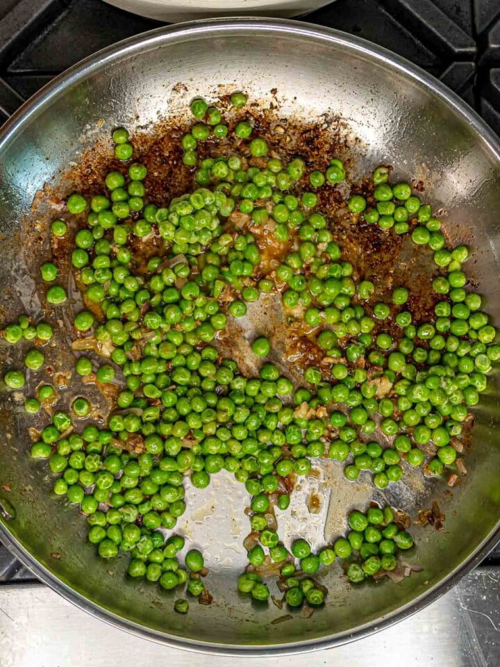 Bright green peas cooking in a stainless steel skillet with browned bits from the sausage.