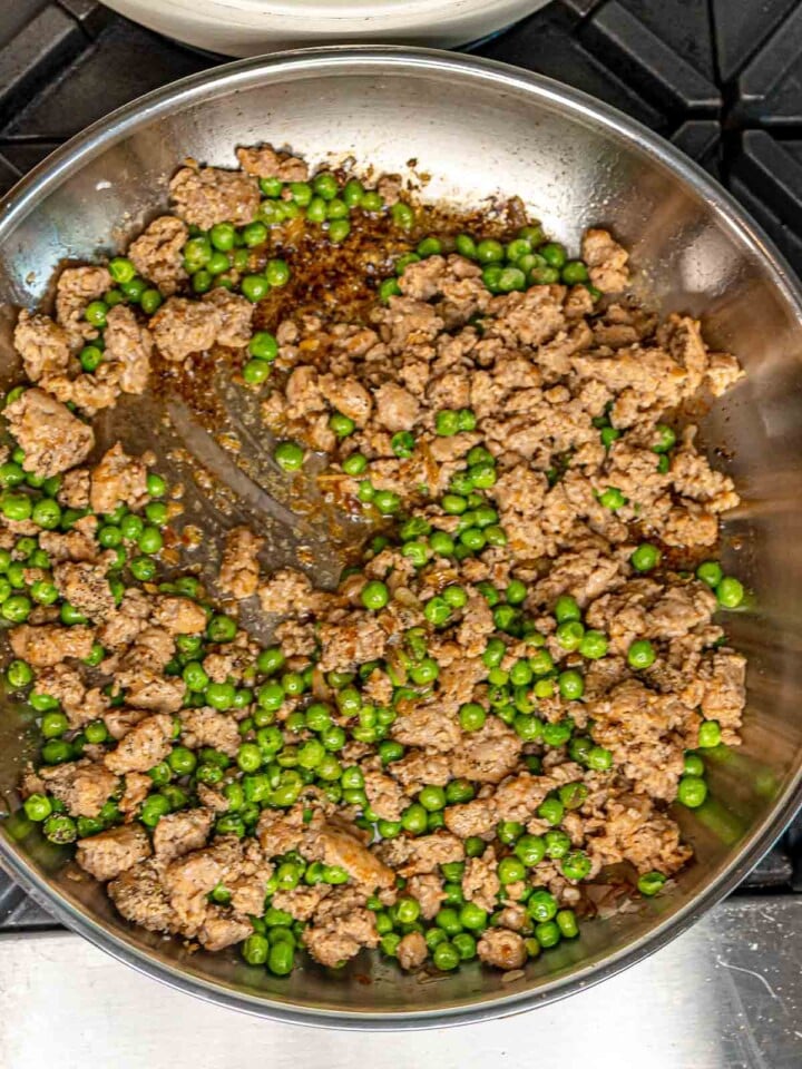 Crumbled sausage and green peas combined in a stainless steel skillet, being stirred with a spatula.