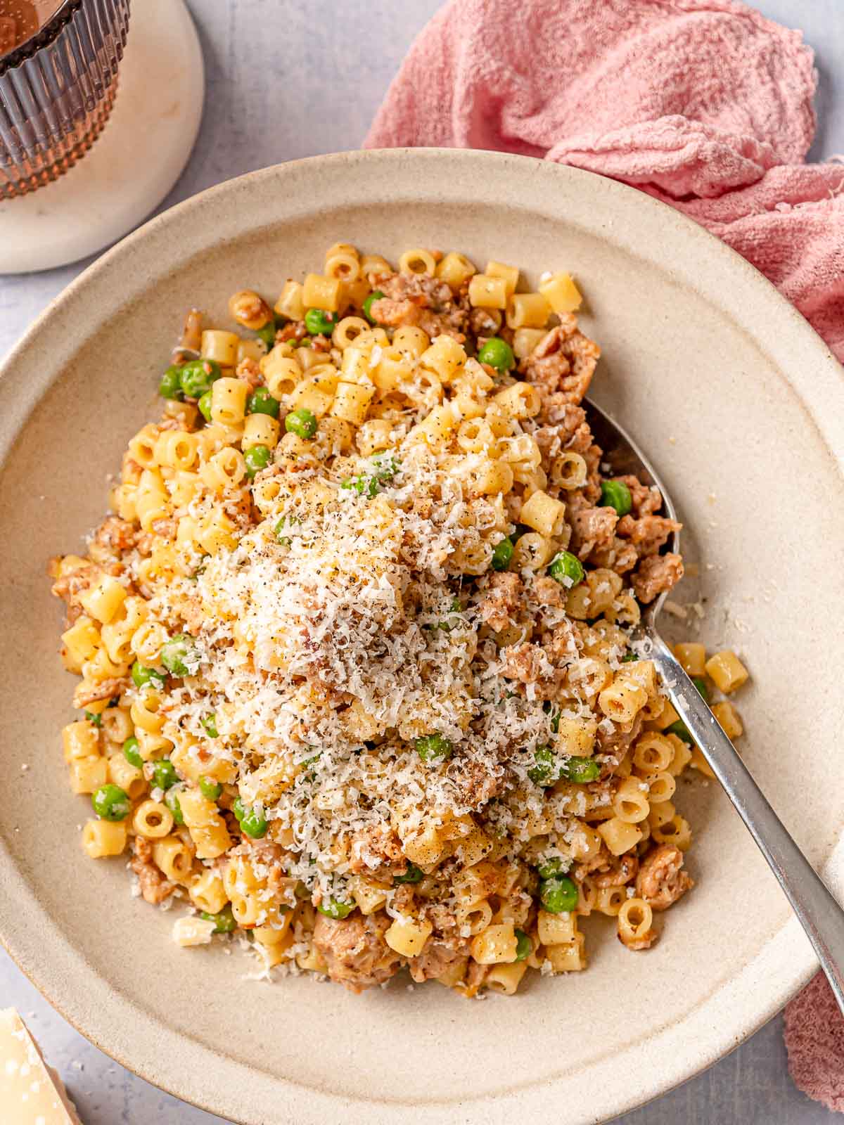 A bowl of ditalini with sausage and peas topped with a generous mound of grated Parmesan and black pepper, with a spoon in the bowl and a pink linen in the background.