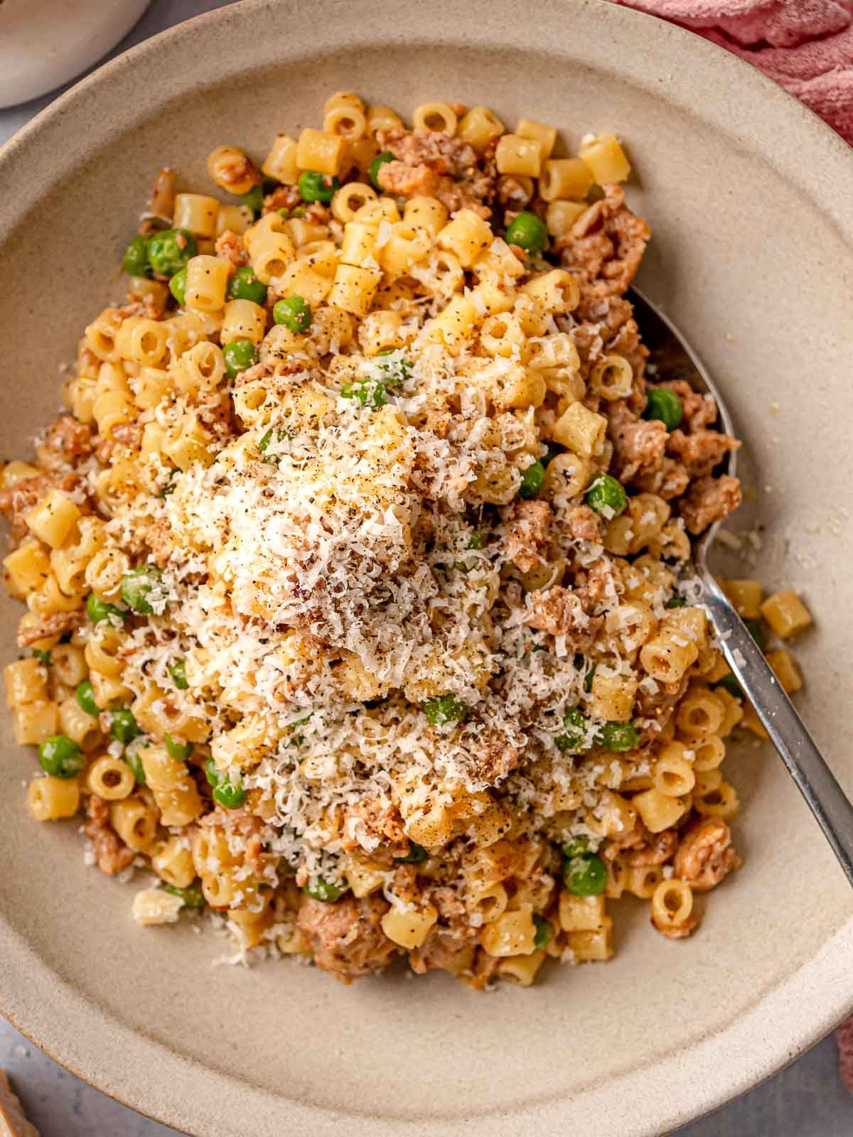 A serving of ditalini with crumbled sausage, peas, and freshly grated Parmesan in a neutral stoneware bowl with a spoon.