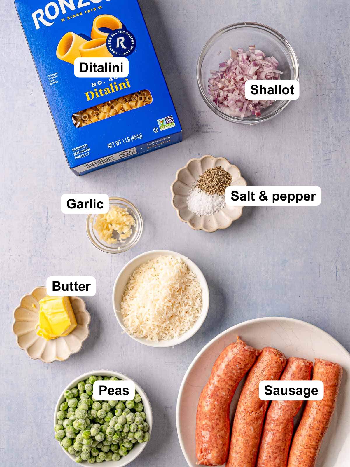 Labeled ingredient layout showing a box of Ronzoni ditalini pasta, diced shallot, minced garlic, salt and pepper, butter, grated Parmesan, frozen peas, and Italian sausage links.