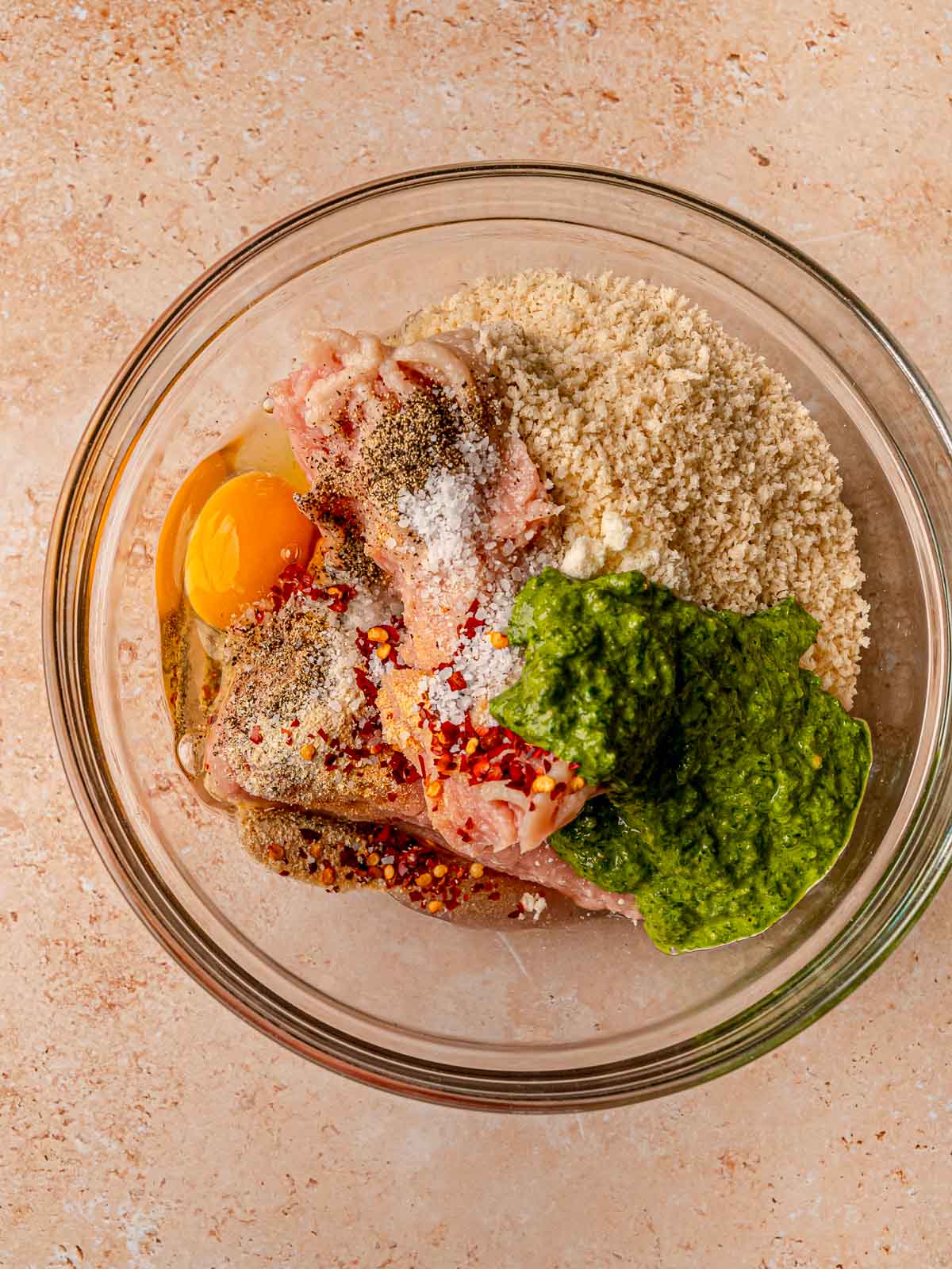 Glass bowl containing ground chicken, egg, breadcrumbs, Parmesan cheese, pesto, and spices before mixing.