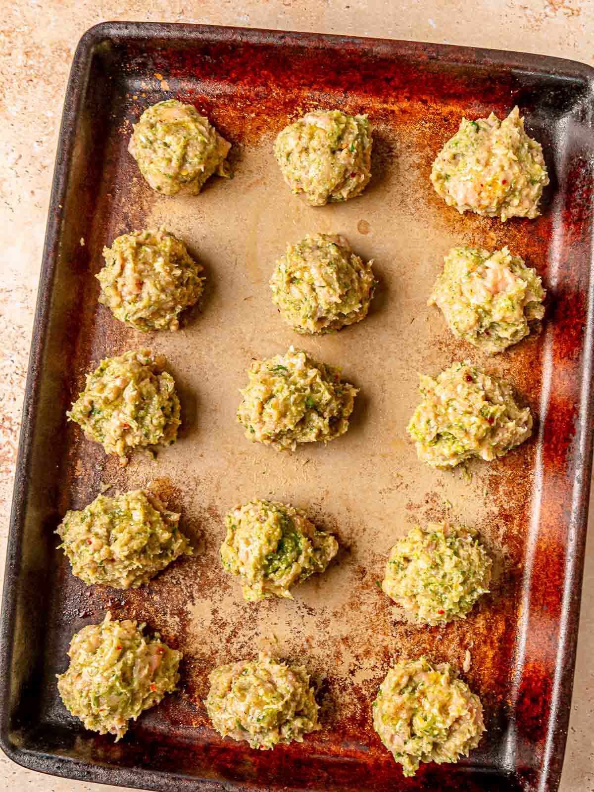 Uncooked pesto chicken meatballs arranged in rows on a dark baking sheet.