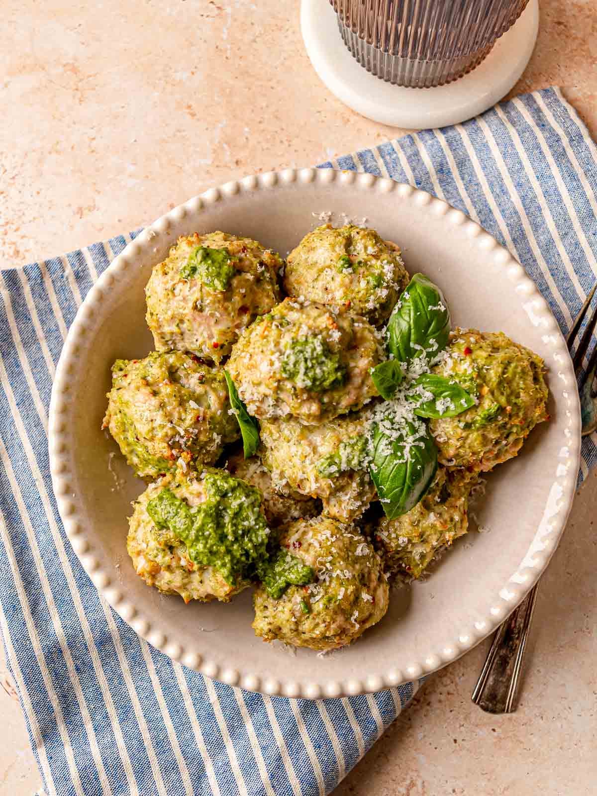 Serving of pesto chicken meatballs on a white scalloped plate with fresh basil and shredded Parmesan cheese on a blue striped kitchen towel.