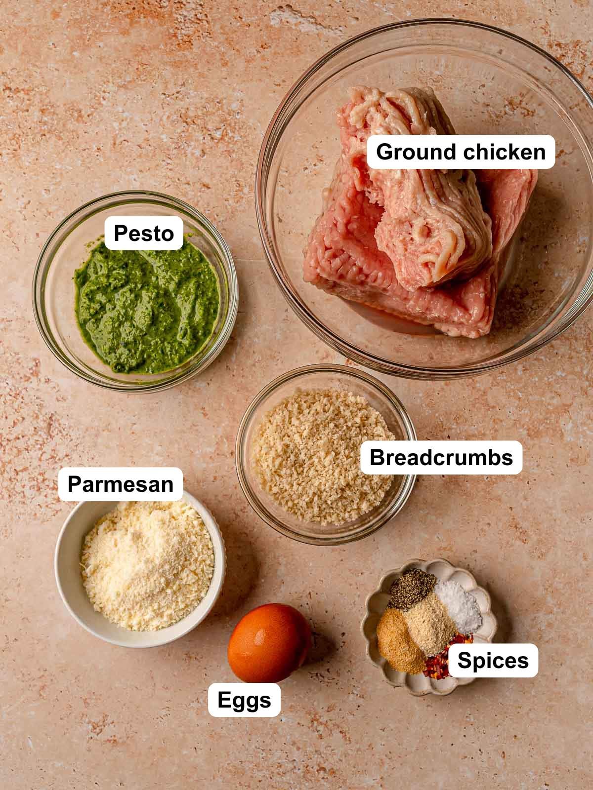 Labeled ingredient bowls showing ground chicken, pesto, breadcrumbs, Parmesan cheese, eggs, and spices.